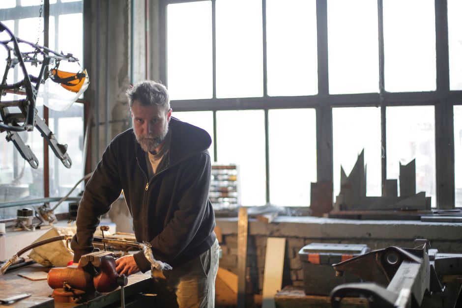 Middle aged bearded workman in workwear standing near workbench and working with equipment in garage and looking at camera